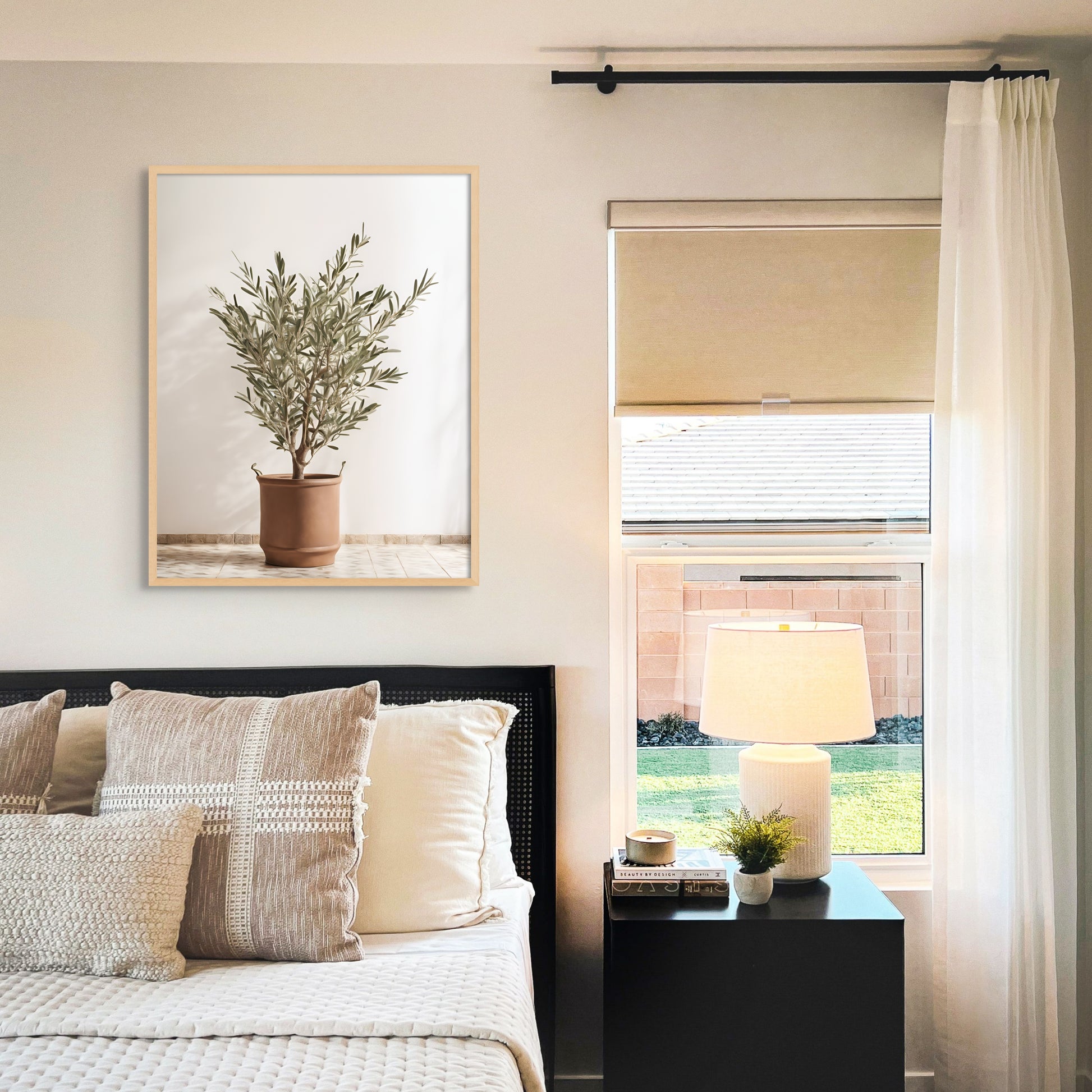 A unique photograph print on canvas of and olive tree in a terracotta color pot with handles. Featuring a bright and sunny background. Displayed over a neutral color bedding.