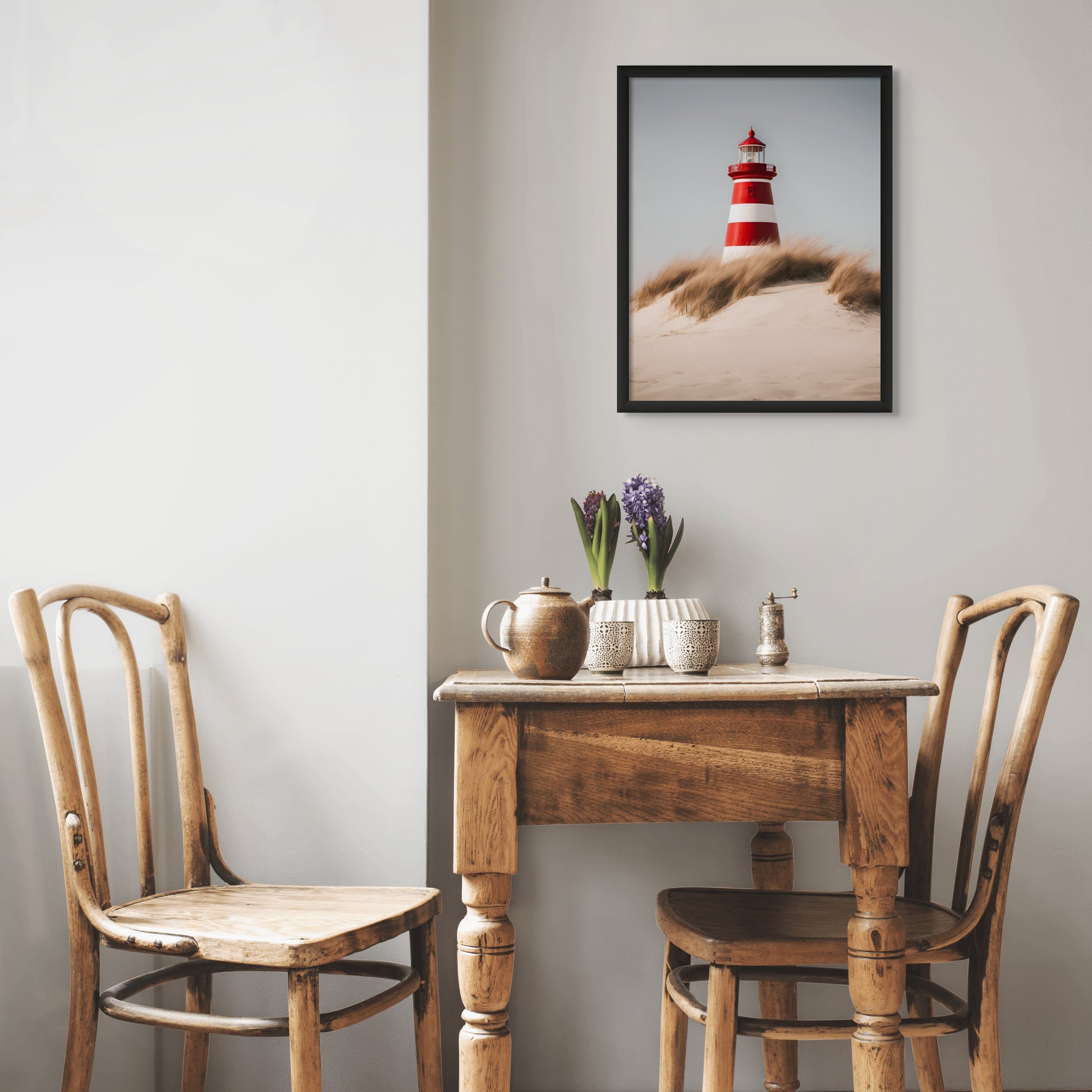a red and white lighthouse from a view in the sand dunes. Featuring wind blown beach grass and a bright sunny day. Displayed above a rusic wood table and chair set