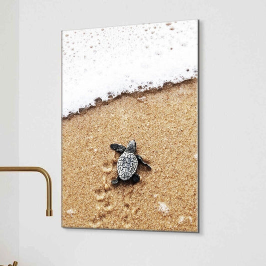 A vertical wall art print showing a baby sea turtle on a sandy beach with white ocean foam, displayed on a light gray wall.